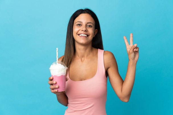 jonge vrouw met aardbei milkshake glimlachen en tonen overwinning teken - Foto, afbeelding
