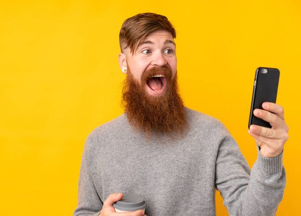 Redhead man with long beard over isolated yellow background holding coffee to take away and a mobile - Foto, Bild