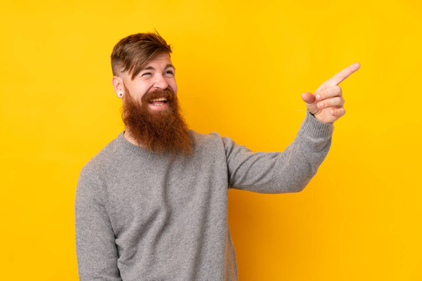 Rotschopf mit langem Bart auf gelbem Hintergrund zeigt mit dem Finger zur Seite und präsentiert ein Produkt - Foto, Bild