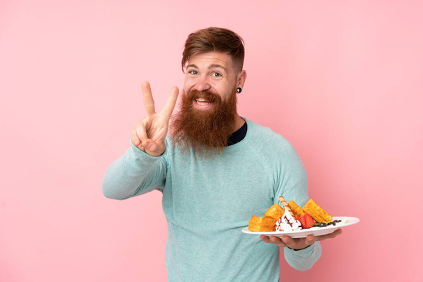 Redhead man with long beard holding waffles over isolated pink background smiling and showing victory sign - Foto, Bild