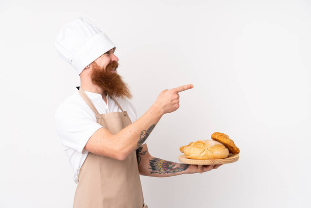 Punapää keittiöpuvussa. Urosleipuri, jolla on pöytä, jossa on useita leipiä, jotka osoittavat sivulle esittämään tuotetta - Valokuva, kuva