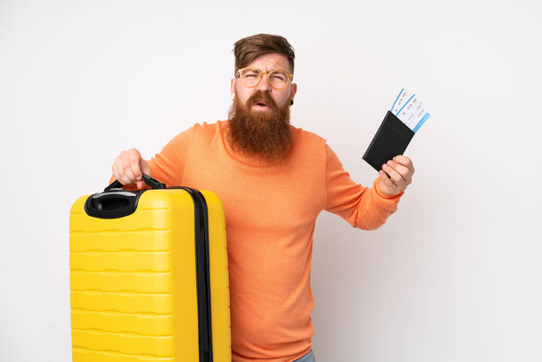 Pelirrojo con barba larga sobre fondo blanco aislado infeliz en vacaciones con maleta y pasaporte - Foto, Imagen