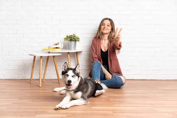 Молодая девушка со своей собакой сидит на полу в помещении улыбаясь и показывая знак победы - Фото, изображение