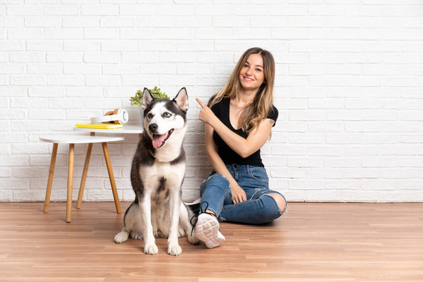 Молодая красивая женщина со своей хаски собака сидит на полу в помещении, указывая на сторону, чтобы представить продукт - Фото, изображение