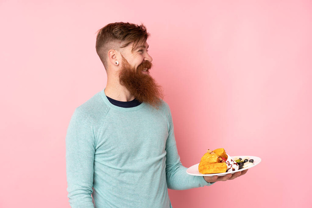 Redhead man with long beard holding waffles over isolated pink background with happy expression - Fotó, kép