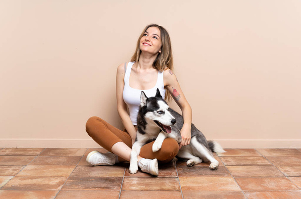 Young pretty woman with her husky dog sitting in the floor at indoors looking up while smiling - Photo, Image