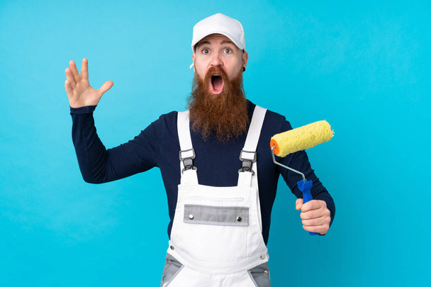 Homem pintor com barba longa sobre fundo azul isolado com expressão facial surpresa - Foto, Imagem