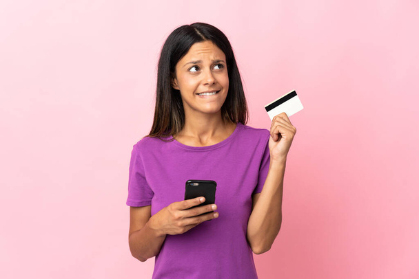 blank meisje geïsoleerd op roze achtergrond kopen met de mobiele telefoon met een creditcard terwijl denken - Foto, afbeelding