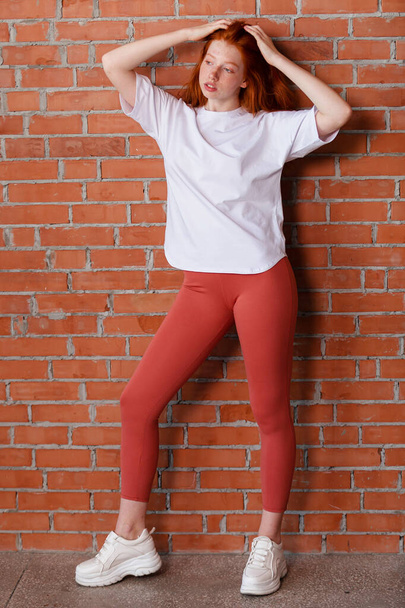 Studio ve stylu módy portrét krásné rusovlasé mladé dívky v legíny kalhoty a bílé tričko. Model stojící a pózující vedle cihlové zdi - Fotografie, Obrázek