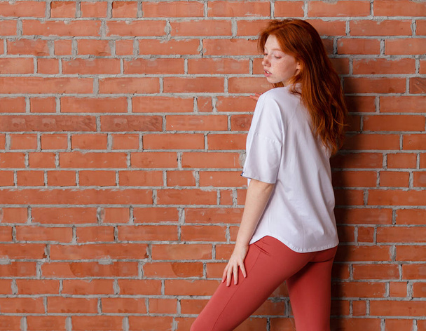 Modestil Studio Porträt der schönen rothaarigen jungen Mädchen in Leggings Hose und weißem T-Shirt. Model steht und posiert neben Mauer - Foto, Bild