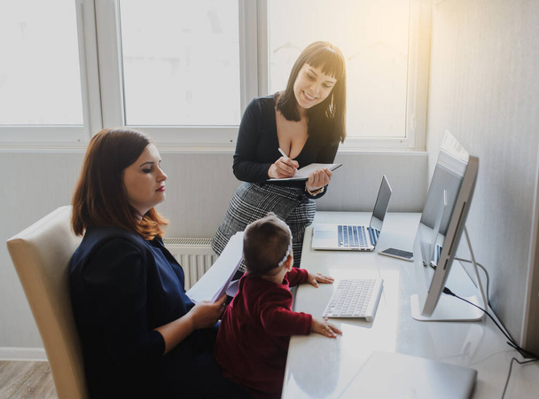 close up photo of two women in office outfit working on computers and discussing a report; one of them is with a baby girl in her arms - Foto, immagini