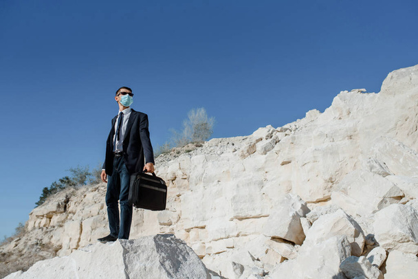 Un giovane uomo d'affari con una valigetta e una maschera per il viso sta scalando la montagna. Concetto di coronavirus COVID 19 crisi pandemica ed economica. - Foto, immagini