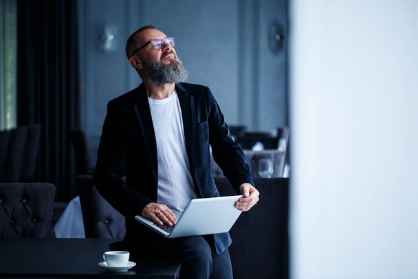 Un homme d'affaires adulte avec une barbe dans les lunettes s'assoit avec un ordinateur portable sur ses genoux et travaille. Le directeur établit un calendrier pour le déroulement du travail - Photo, image