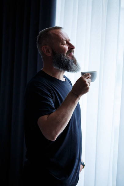 髭を生やした大人の男がコーヒーを飲み、窓から外を見る。作業日フィールドレクリエーションのコンセプト - 写真・画像