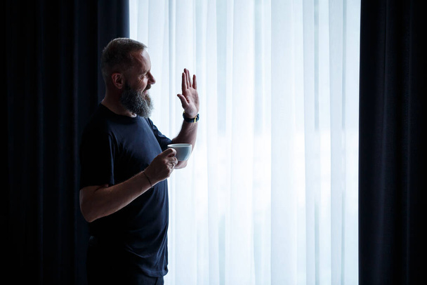 An adult man with a beard drinks coffee and looks out the window. Working day field recreation concept - Photo, Image