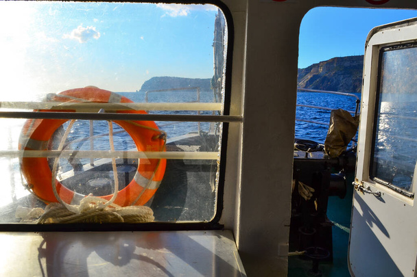 inside cabin of old yacht, ship with windows and red life buoy, white door in the light of sun in blue sea with rocks, mountains - Foto, Bild