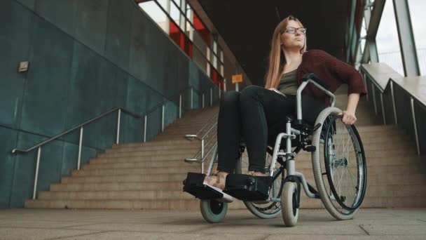 Młoda, zniechęcona biała kobieta na wózku inwalidzkim przed klatką schodową rozglądająca się za dostępną drogą - Materiał filmowy, wideo