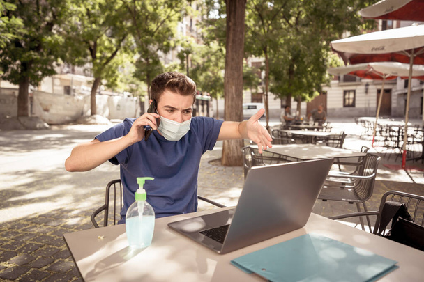 Young man wearing protective face mask and using mobile on video conference working remotely on computer laptop in coffee shop outdoors. The New Normal and health regulations against COVID-19. - Photo, Image