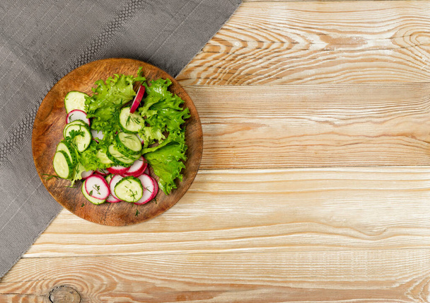 Spring homemade radish salad with fresh cucumber and greens on wooden plate top view. Simple green rustic salat mockup with copy space - Photo, Image