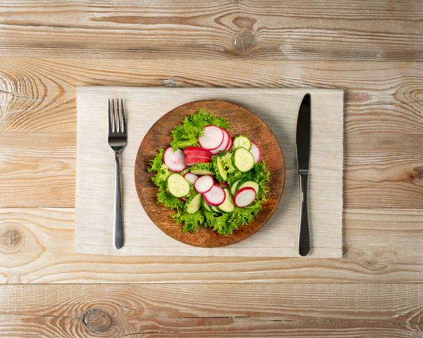 Spring homemade radish salad with fresh cucumber and greens on wooden plate top view. Simple green rustic salat with sliced radishes, cucumbers, escarole and lettuce - Photo, Image
