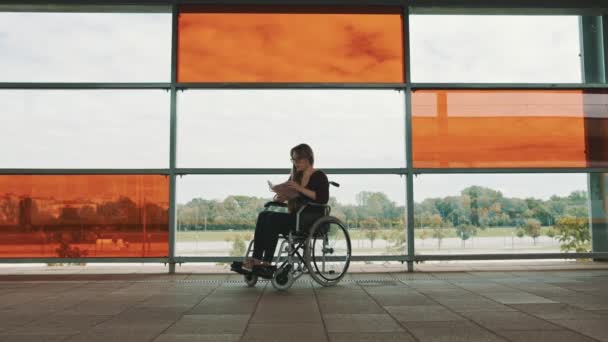 Young independed caucasian woman in the wheeldchair reading a book in front of big glass multicoloroed windows - Footage, Video