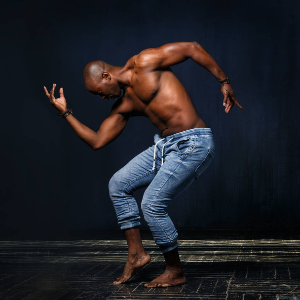 A black athletic man in jeans with a naked torso is dancing against a dark background. - Фото, зображення