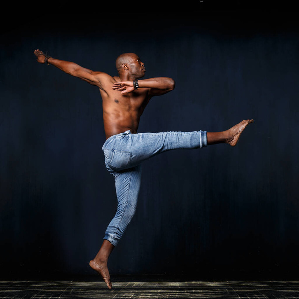 A black athletic man in jeans with a naked torso is dancing against a dark background. - Photo, image
