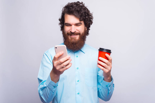 Ένας άντρας κοιτάζει και χαμογελάει στο τηλέφωνό του και κρατάει ένα φλιτζάνι με ζεστό ποτό. - Φωτογραφία, εικόνα
