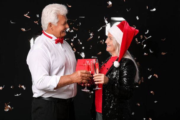 Elderly couple with champagne and Christmas gift on dark background - Фото, зображення