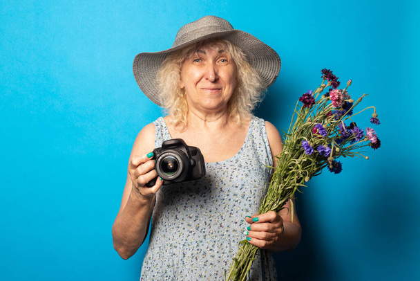 Старуха в широкополой шляпе и платье с букетом цветов и фотоаппаратом на синем фоне. - Фото, изображение