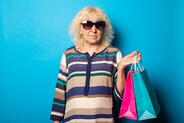 Gülümseyen yaşlı kadın mavi arka planda alışveriş torbaları tutuyor.. - Fotoğraf, Görsel
