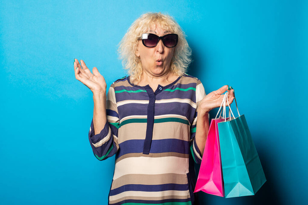 Şaşkın suratlı yaşlı kadın mavi arka planda alışveriş torbaları tutuyor.. - Fotoğraf, Görsel