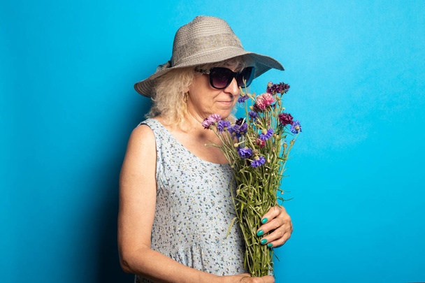Old woman in a wide-brimmed hat and dress sniffing a bouquet of flowers on a blue background. - Фото, изображение