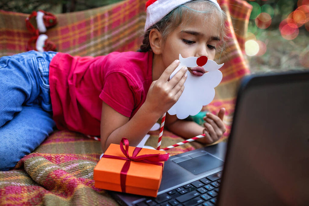 Nová normální online vánoční oslava. Roztomilý sourozenec pomocí notebooku a slavnostní zábavné rekvizity na oslavu Vánoc s rodinou přes video chat, šťastné svátky, venkovní - Fotografie, Obrázek