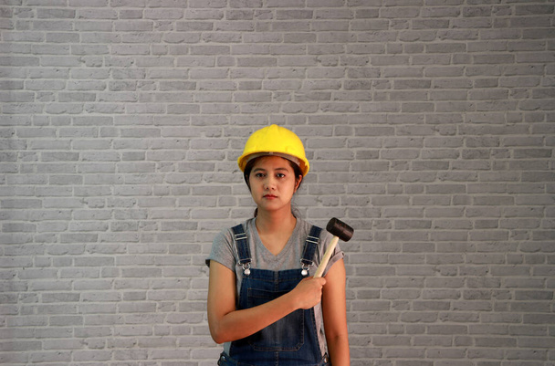 Technicien femme ware casque jaune avec T-shirt gris et jeans denim robe tablier debout et marteau en acier à la main sur fond de motif en brique grise. - Photo, image