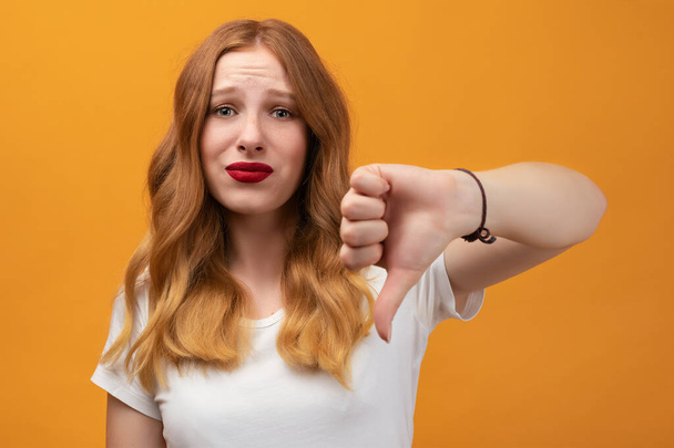 波状の赤毛のかなり若い女性は、親指を示す白いTシャツを着ている。黄色の背景に隔離された、嫌い、失敗ジェスチャーの概念とは異なり、 - 写真・画像