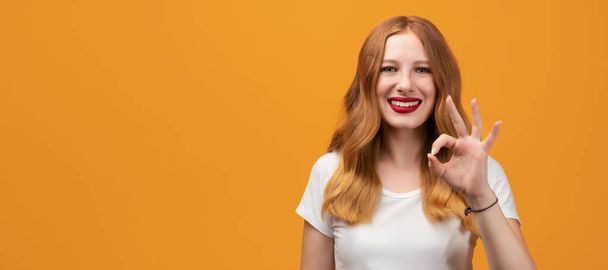 Happy girl with long wavy redhead, wearing white t-shirt Smiling and showing OK gesture. I like that. Good job. Human emotions concept. Copy space for your text - Photo, Image