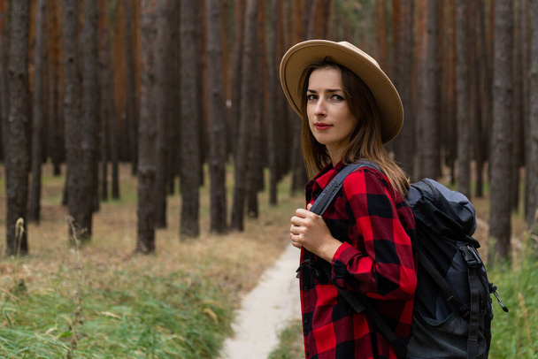 Młoda kobieta w kapeluszu, czerwonej koszuli i plecaku w lesie. Wędrówka po lesie. - Zdjęcie, obraz