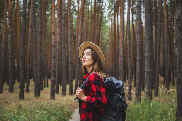 Young woman in a hat, red shirt and backpack looks at the treetops in a pine forest. Camping in the woods. - Фото, изображение