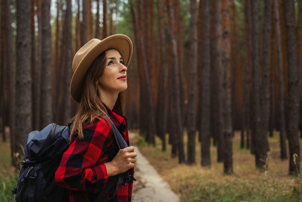 Młoda kobieta w kapeluszu, czerwonej koszuli i plecaku patrzy na wierzchołki drzew w sosnowym lesie. Camping w lesie. - Zdjęcie, obraz