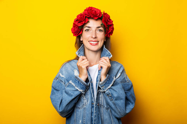 若いです女性で笑顔でデニムジャケットと赤い花の花輪で彼女の頭の上に黄色の背景. - 写真・画像