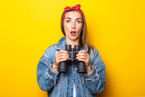 Young woman with a surprised face in jeans and a raft on her head holds binoculars in her hands on a yellow background. Banner. - Photo, Image