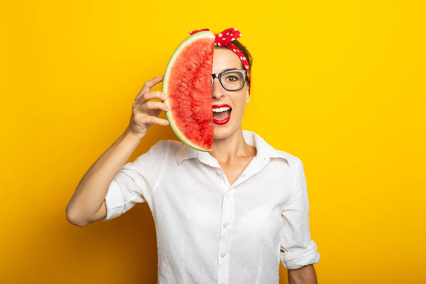 Young woman in a red headband and glasses covers half of her face with watermelon on a yellow background. - Foto, immagini