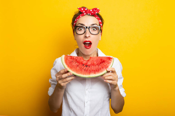 Junge Frau mit überraschtem Gesicht, rotem Stirnband und Brille isst eine Wassermelone auf gelbem Hintergrund. - Foto, Bild