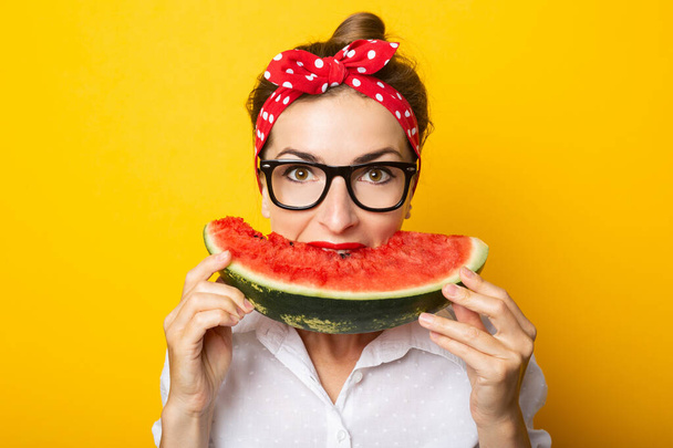 Young woman with a smile in a red headband and glasses eats watermelon on a yellow background. - Foto, afbeelding