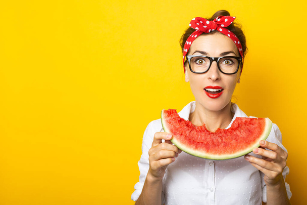 Młoda kobieta z uśmiechem w czerwonej opasce i okularach zjada arbuza na żółtym tle. - Zdjęcie, obraz