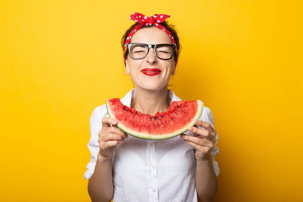 Молода жінка з посмішкою в червоній смузі голови і окулярах їсть кавун на жовтому тлі
. - Фото, зображення