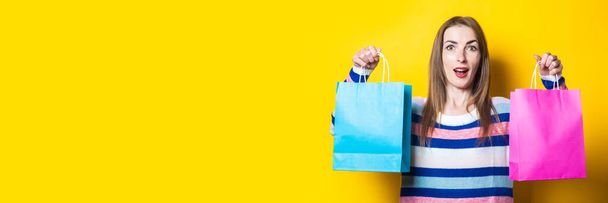 Mujer joven con una sonrisa en un suéter sostiene bolsas de compras con compras en un fondo amarillo. Banner. - Foto, Imagen
