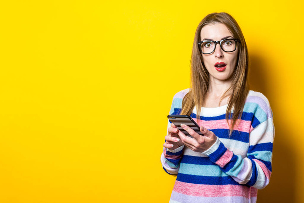 Young woman with a surprised face and holding a phone on a yellow background. Banner. - Fotoğraf, Görsel