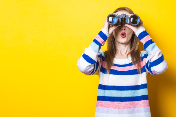 Young woman with a surprised face looks through binoculars on a yellow background. Banner. - Zdjęcie, obraz